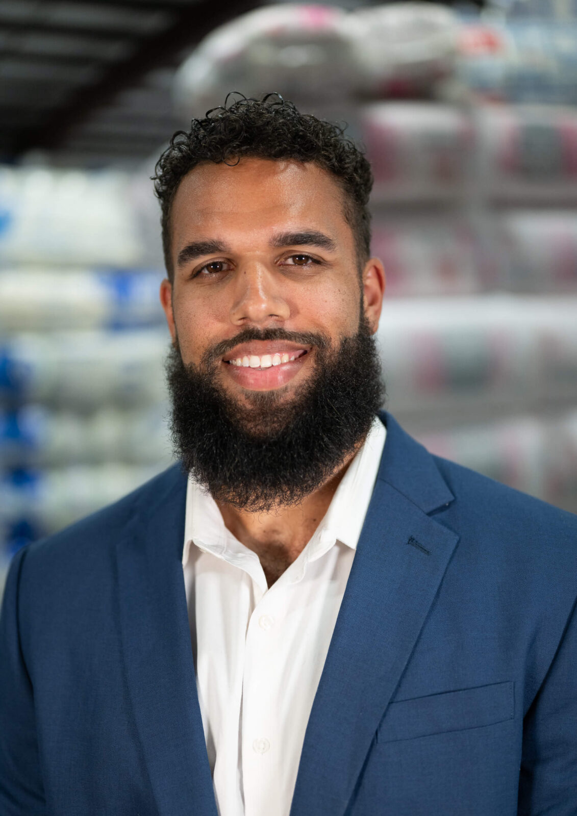 Nick Hughes Smiling man with a beard in a warehouse, wearing a blue suit and white shirt, background blurred with insulation rolls