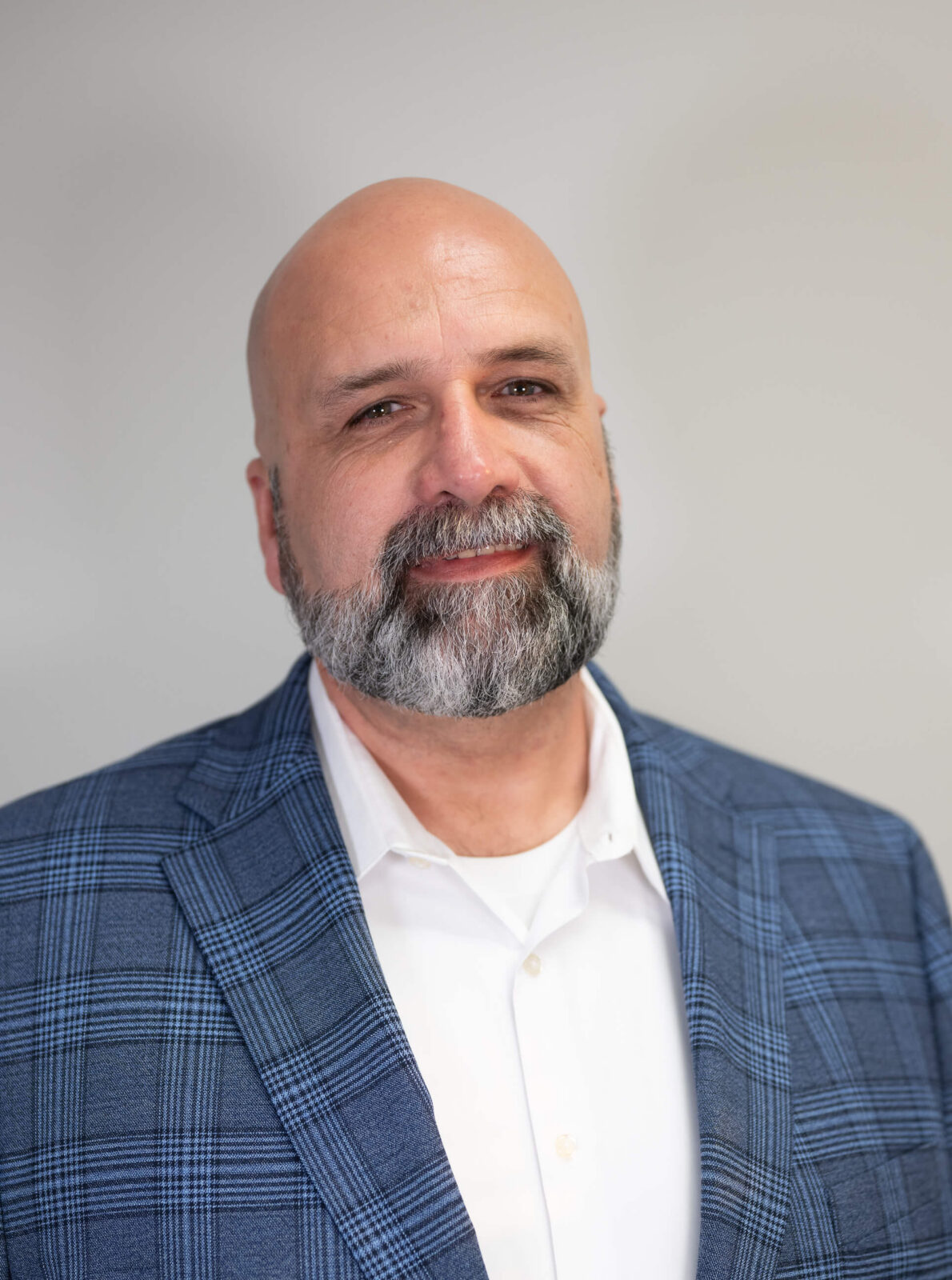 A man with a bald head and a beard wearing a blue plaid blazer and white shirt, smiling slightly against a gray background